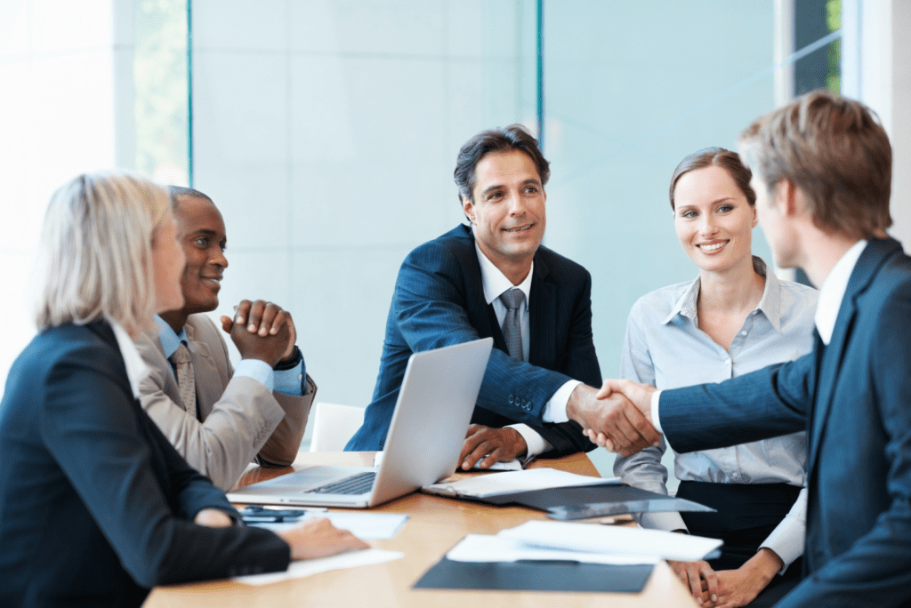 Team di lavoro seduto alla scrivania con computer portatili, mentre due membri del team si stringono la mano in segno di accordo o collaborazione, in un ambiente professionale e dinamico.