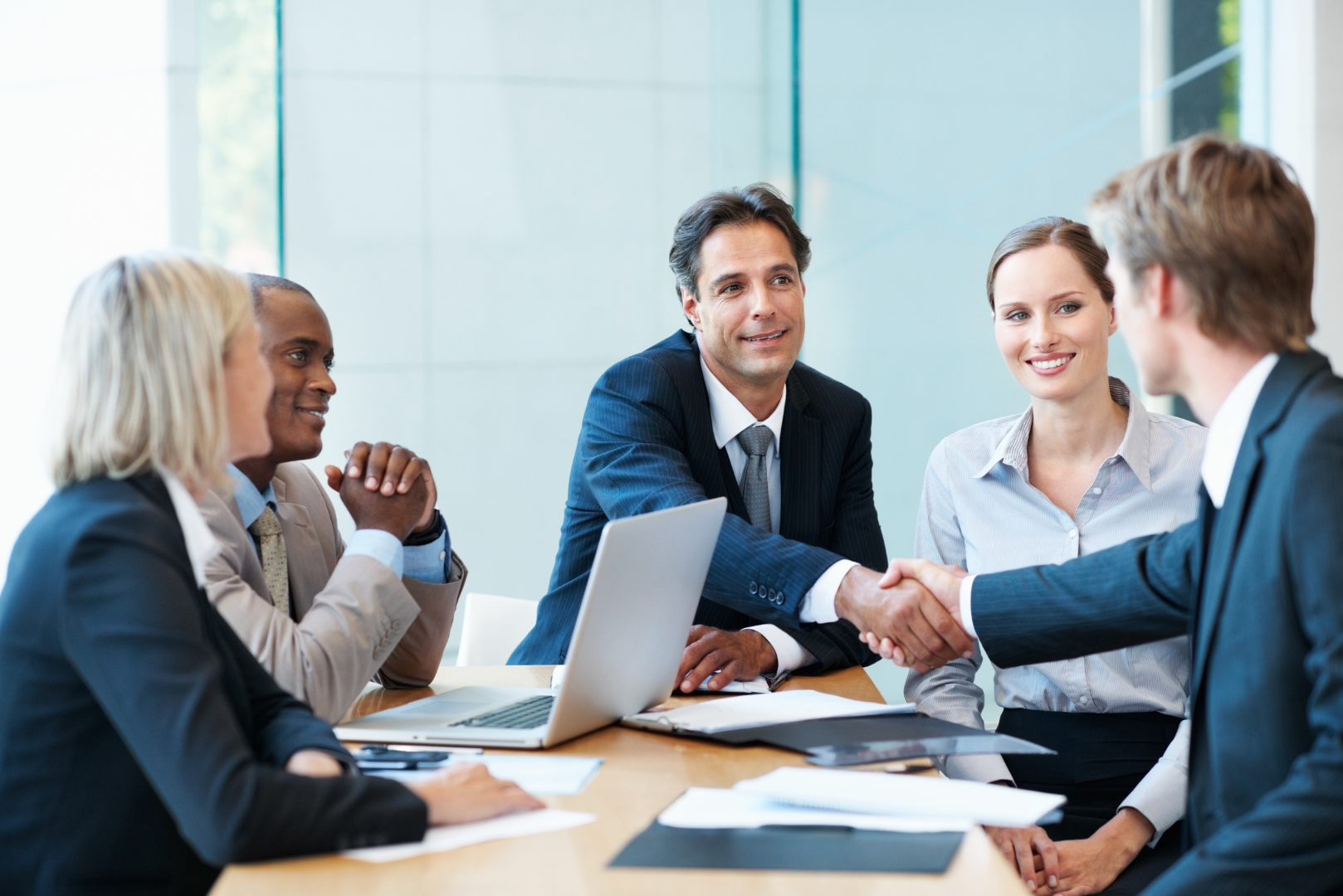 Team di lavoro seduto alla scrivania con computer portatili, mentre due membri del team si stringono la mano in segno di accordo o collaborazione, in un ambiente professionale e dinamico.