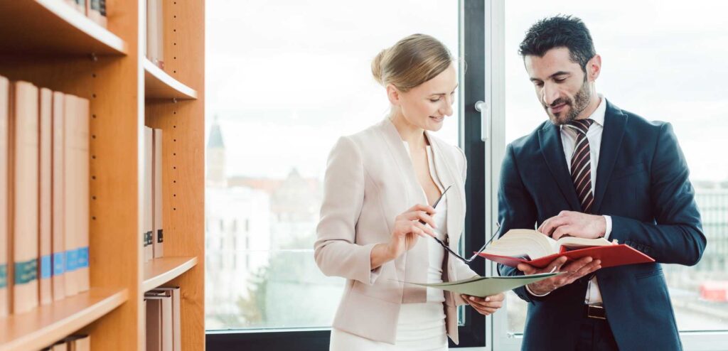 Donna e uomo, entrambi in abbigliamento professionale, parlano tra loro mentre consultano dei libri, scambiandosi idee o informazioni in un ambiente di lavoro collaborativo.