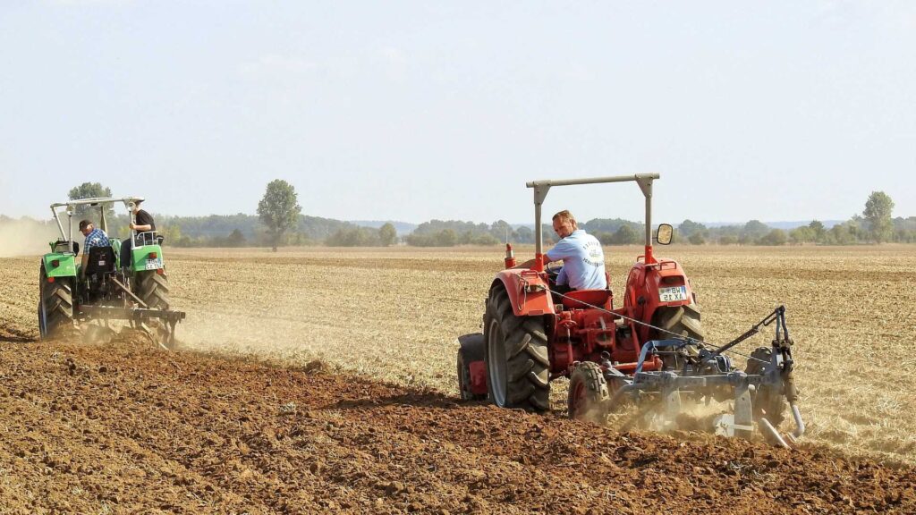 Trattori in azione mentre arano un ampio campo, con il terreno lavorato sotto le ruote e l'ambiente rurale circostante, in un paesaggio agricolo.