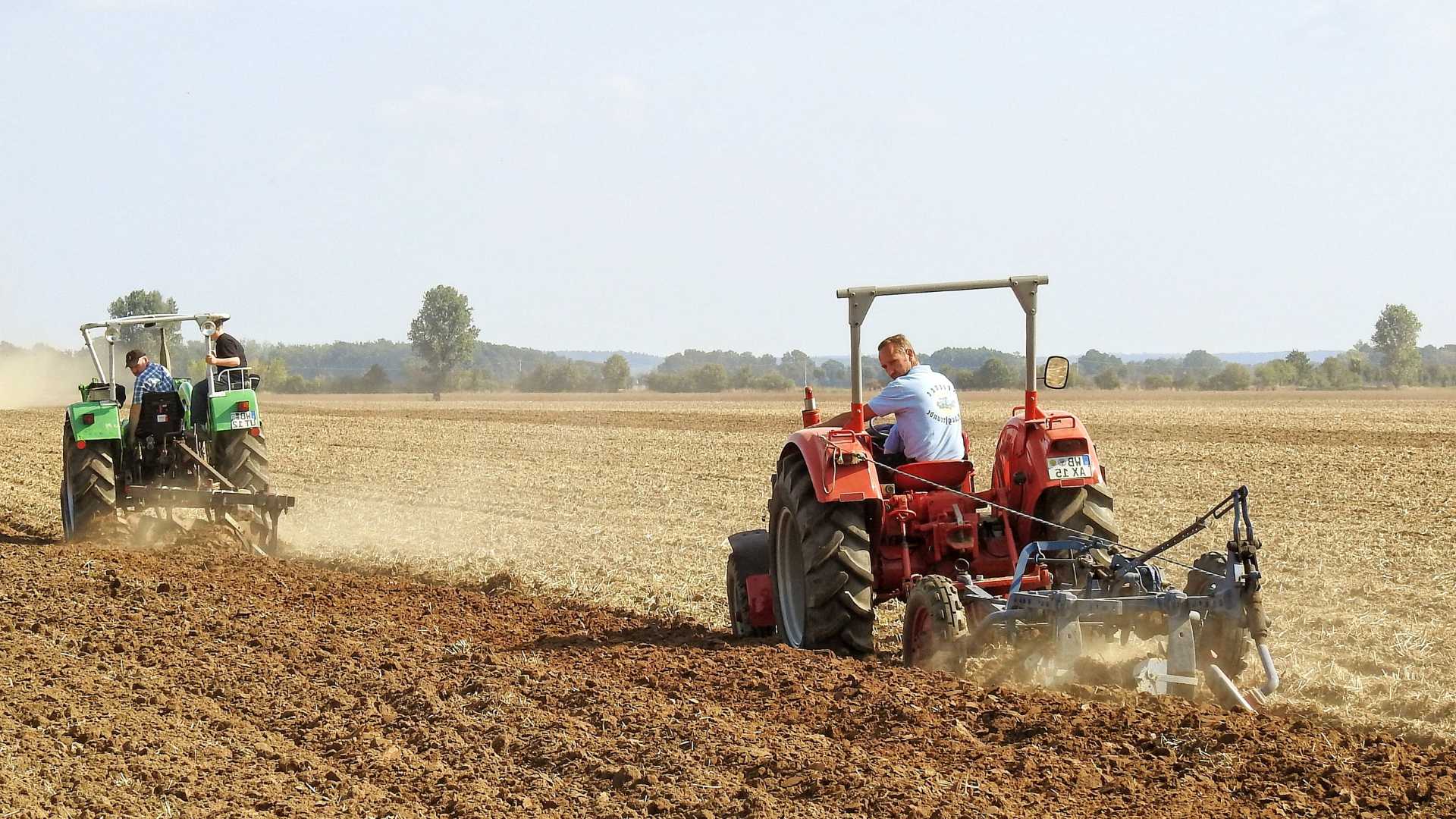 Trattori in azione mentre arano un ampio campo, con il terreno lavorato sotto le ruote e l'ambiente rurale circostante, in un paesaggio agricolo.