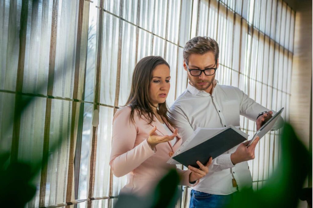 Un uomo e una donna, entrambi in abbigliamento professionale, discutono di lavoro mentre esaminano dei documenti su una scrivania, mostrando attenzione e collaborazione in un ambiente di ufficio.