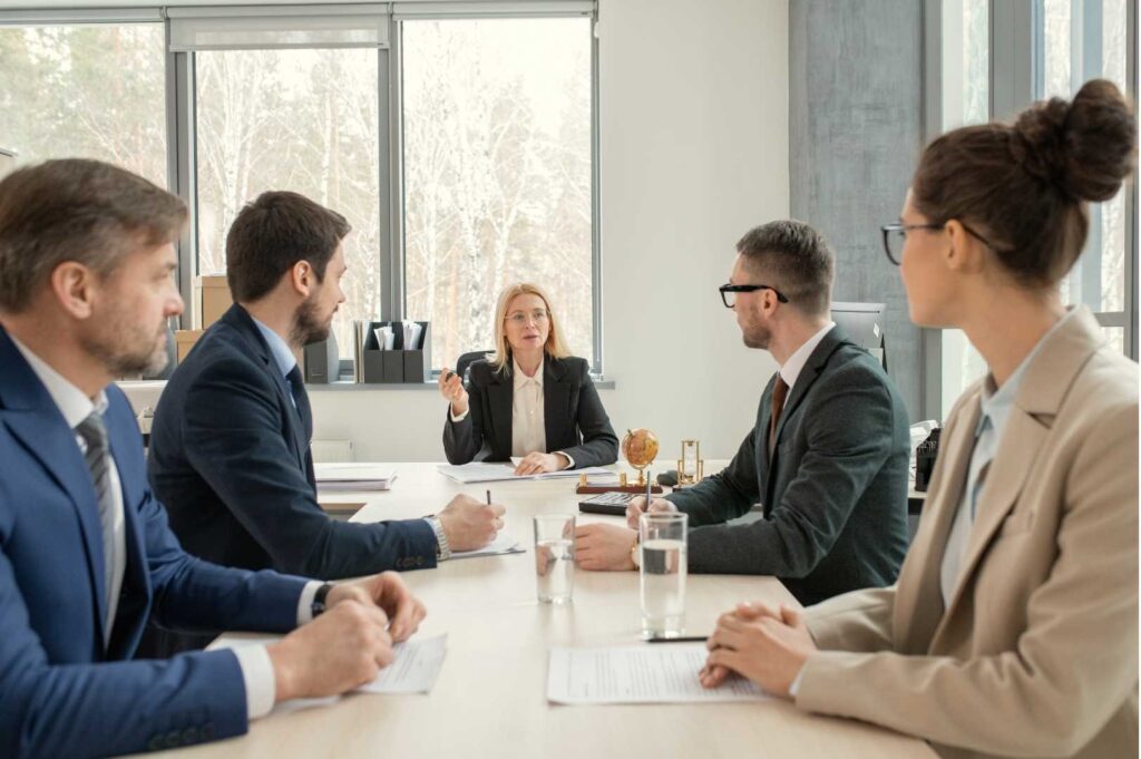 Consulenti in abbigliamento professionale parlano tra loro attorno a una scrivania quadrata, scambiandosi idee e lavorando in team in un ambiente di ufficio moderno.