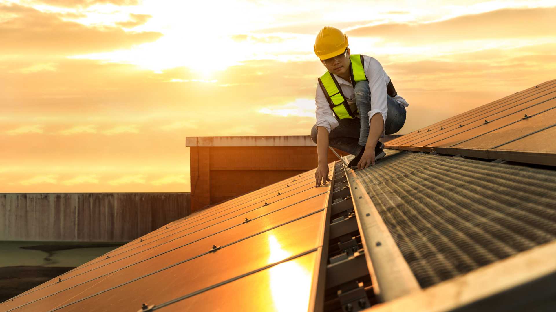Operaio al lavoro su un tetto, mentre installa un pannello solare, indossando attrezzature di sicurezza e lavorando in un ambiente all'aperto, con il panorama circostante visibile.