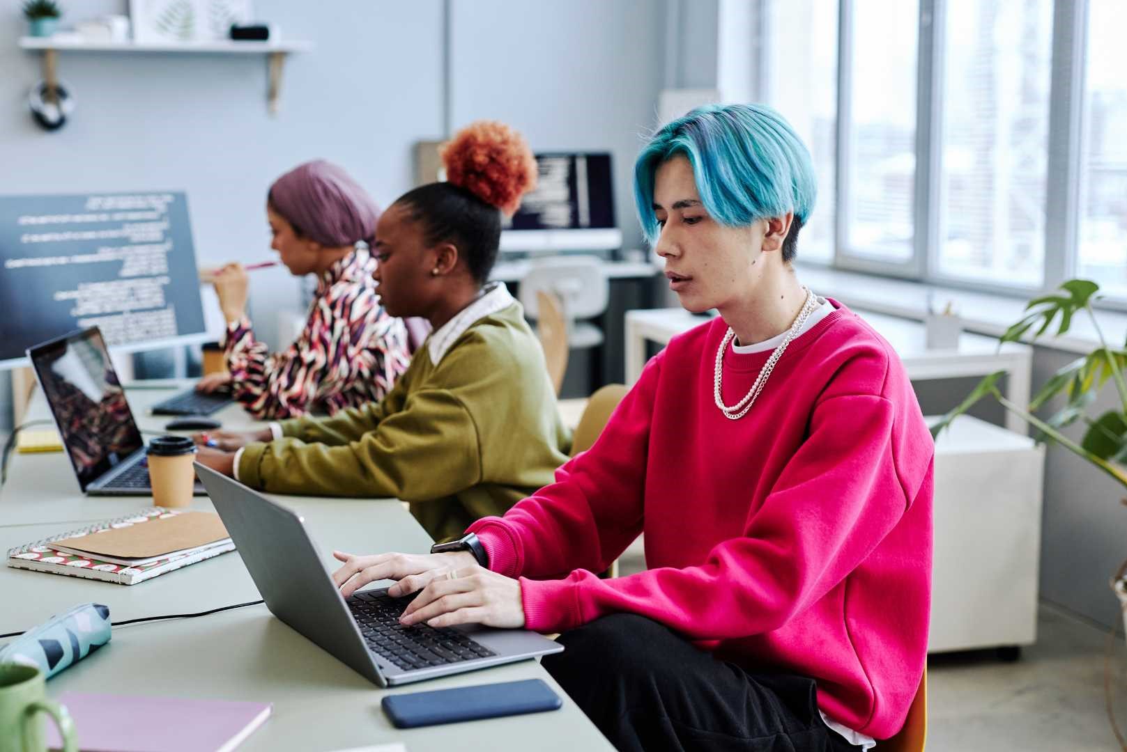 Giovani ragazzi e ragazze con felpe colorate lavorano al computer, concentrati su un progetto in un ambiente informale e creativo.