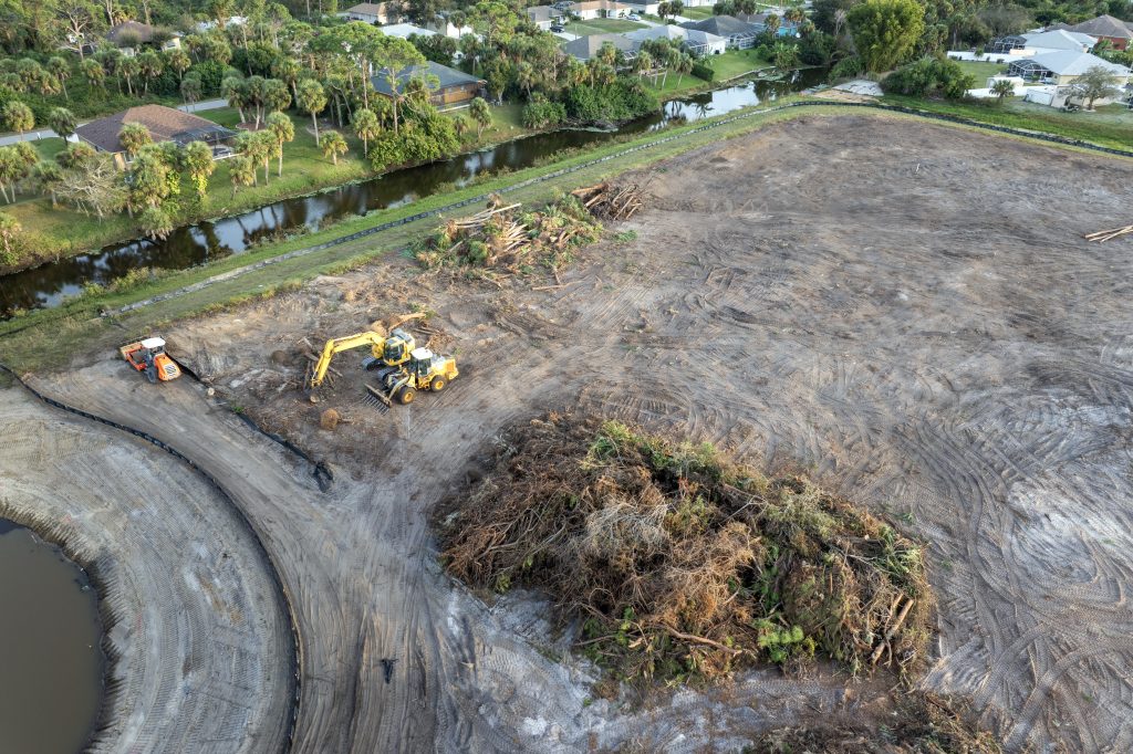 Aerial view of a cleared construction site with heavy machinery, vegetation piles, a canal, and nearby homes.